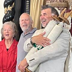 Rabbi Marc holds a Torah with a joyful smile on his face. A man and a woman stand next to him, smiling and singing.
